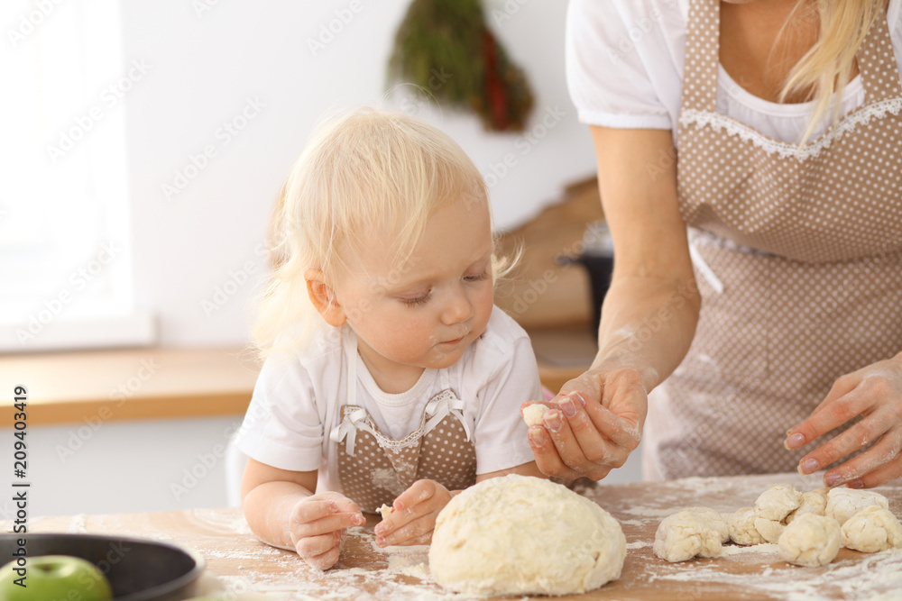 Little girl and her blonde mom in beige aprons  playing and laughing while kneading the dough in the kitchen. Homemade pastry for bread, pizza or bake cookies