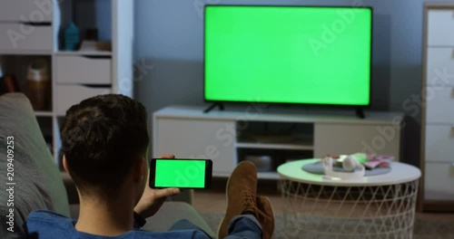 View from behind on the man sitting in the living room in front of the TV with green screen, taping and watching something on the black smartphone with green screen horizontally. Chroma key. Inside