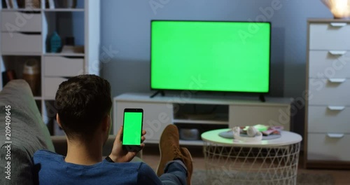 back view on the man sitting in the living room in front of the TV with green screen, taping and watching something on the black smartphone with green screen vertically. Chroma key. Indoor