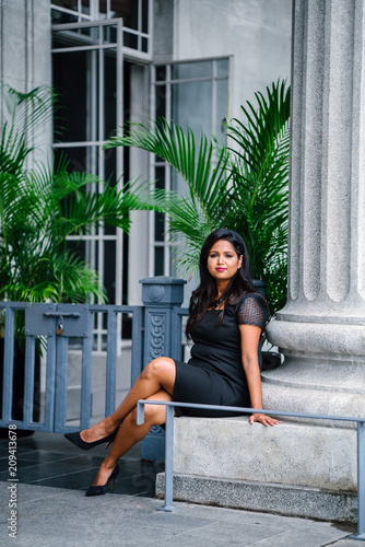 An image of a successful and attractive Indian Asian woman smiling and while sitting and striking a glamorous pose. She is leaning on aconcrete pillar and she is wearing an elegant black dress during  photo