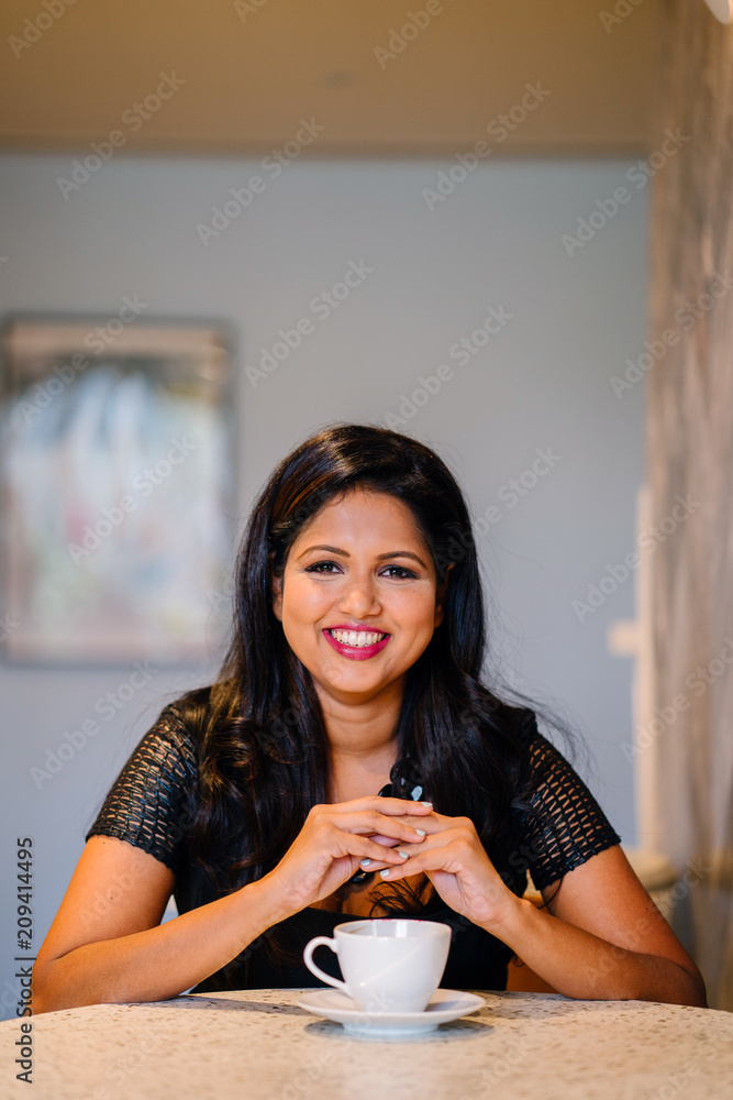 Portrait of a youthful but confident and mature Indian woman smiling while patiently waiting for her colleagues. She is wearing a professional black dress and having a cup of coffee. Portrait of a youthful but confident and mature Indian woman smiling while patiently waiting for her colleagues. She is wearing a professional black dress and having a cup of coffee.