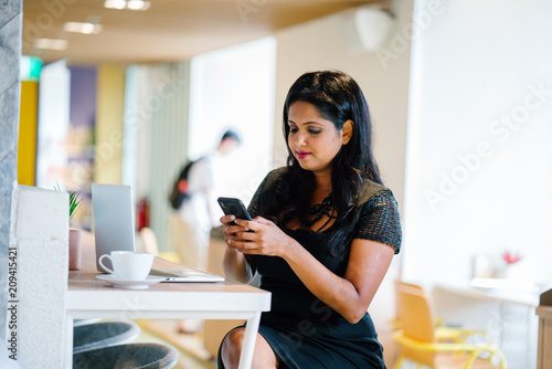 Portrait of a young Indian Asian business woman checking her phone with her laptop computer in front of her. .She is seated in a cafe during the day.She is smiling as she read on the messages she have