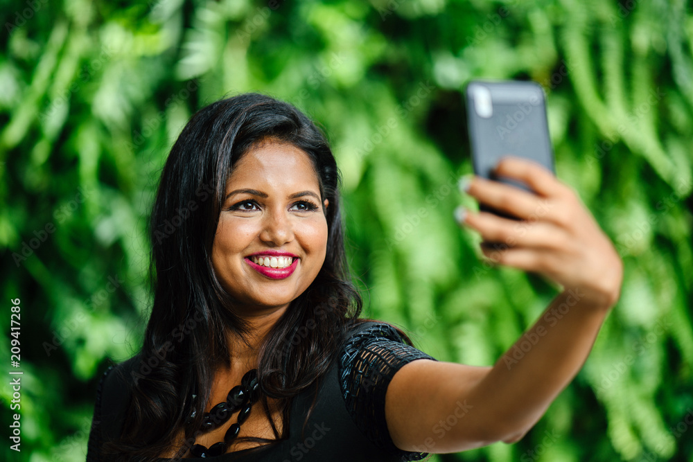 An attractive and professionally dressed Indian Asian woman takes a selfie of herself in the day against a green wall of plants.