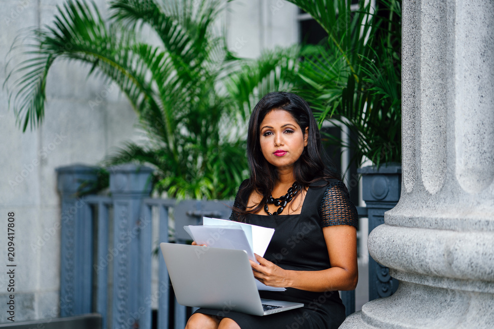 A mature and confident Indian Asian woman sits on the steps of a legal looking building and is working on her laptop computer with some documents. A mature and confident Indian Asian woman sits on the steps of a legal looking building and is working on her laptop computer with some documents.