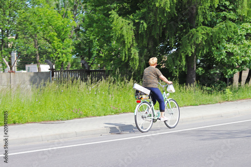 A woman rides a bicycle on the side of the highway against a background of green trees on a sunny, clear day. Alternative Masow type of daily transport. A healthy and athletic way of life. Ecological 