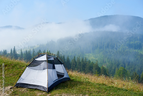 Summer view of small comfortable modern white and blue tourist tent on grassy hill under beautiful blue sky on foggy mountains covered with forest background. Camping, tourism and traveling concept.