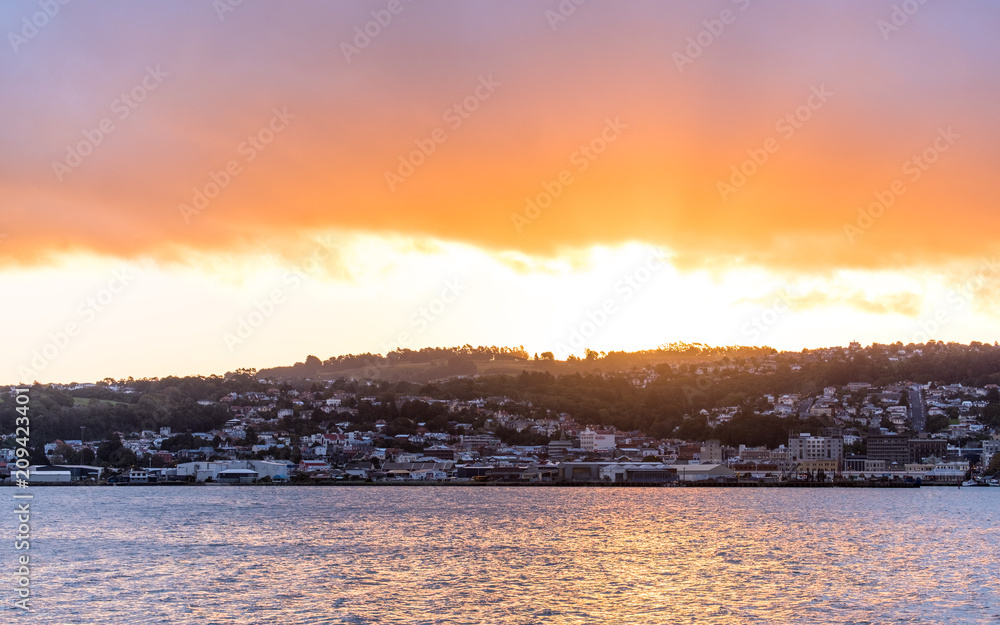 An explosion of sunset light rays over the city and mountain with reflection on the sea. Dunedine, New Zealand.