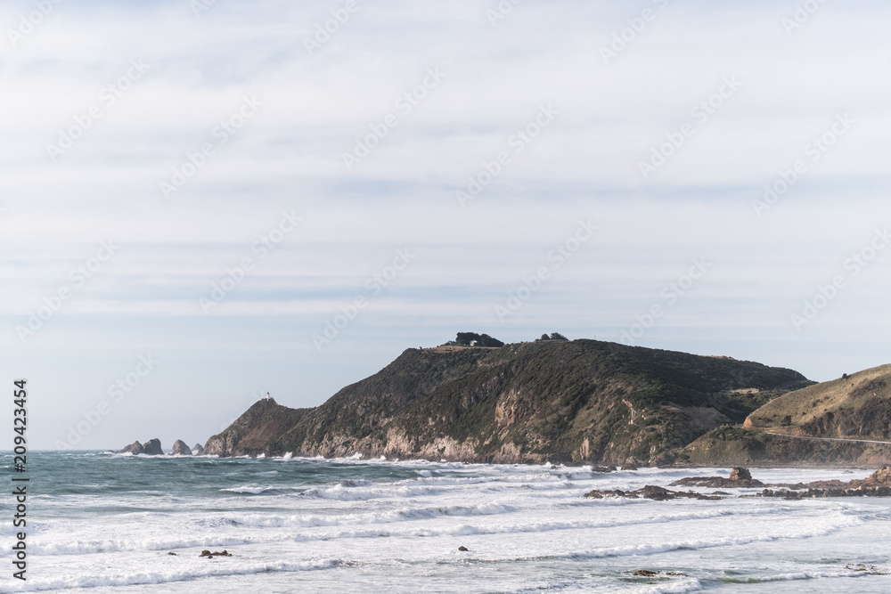 A scene of a beach on a cloudy day with mountain and road to the lighthouse, Nugget point, the Catlins, New Zealand