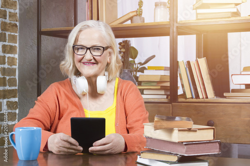 Progressive modern granny concept. Elderly woman wearing colorful clothes with headphones and glasses, smiling, holding tablet / book reader and sitting at the table with blue cup and heap of books