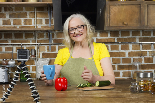 Progressive modern granny concept. Elderly woman blogger wearing glasses, yellow t-shirt and green apron recording her cooking by video camera, drinking tea / coffee from blue cup in kitchen