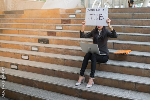 business woman unemployed from company sitting on the staircase and holding poster with text is looking for job, concept of unemployment problem