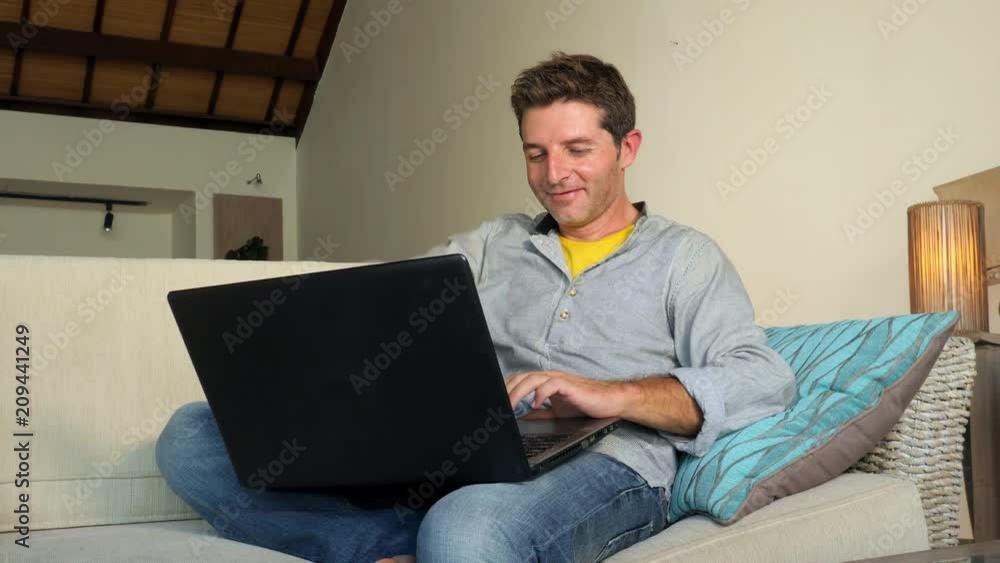 lateral pan on young attractive happy man at home relaxed working on laptop computer sitting at living room sofa couch smiling cheerful in independent entrepreneur and successful freelance job concept lateral pan on young attractive happy man at home relaxed working on laptop computer sitting at living room sofa couch smiling cheerful in independent entrepreneur and successful freelance job concept