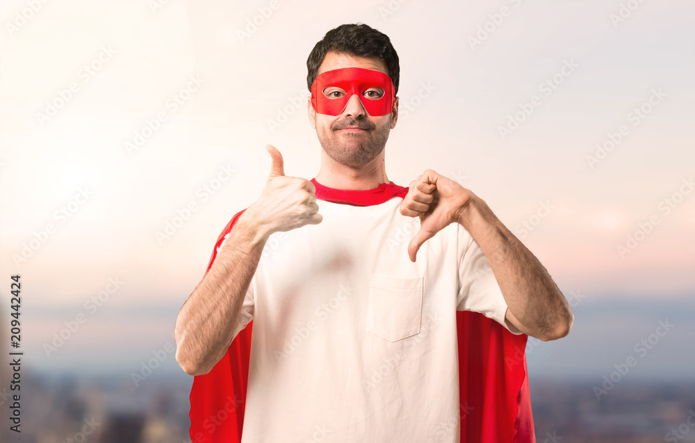 Naklejka premium Superhero man with mask and red cape making good-bad sign. Undecided person between yes or not on a sunset background