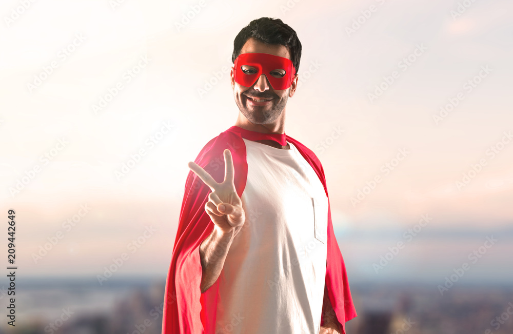 Superhero man with mask and red cape making victory gesture on a sunset background Superhero man with mask and red cape making victory gesture on a sunset background