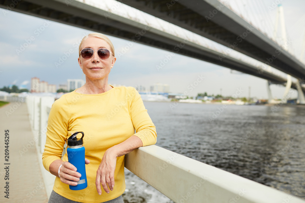 Aged and sporty female with bottle of water leaning at bridge while having rest after running