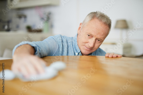 Senior man leaning over table while wiping it with duster during housework