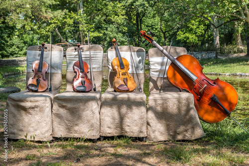 Music and nature concept. String instruments, one cello and three violins on the ceremonial chairs in nature, in front of forest wooden bridge