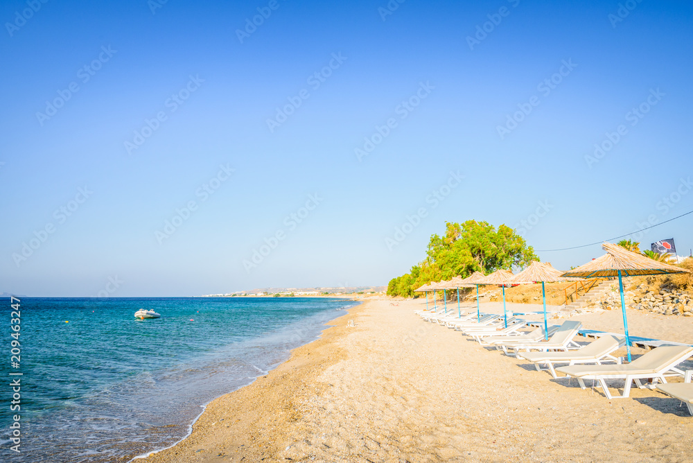 Fototapeta premium Beaches, Greece, Kos Island, Kardamena: beautiful holiday setting on a secluded beach with umbrellas on the Greek Aegean Sea with turquoise waters and a picturesque bay and islands in the background