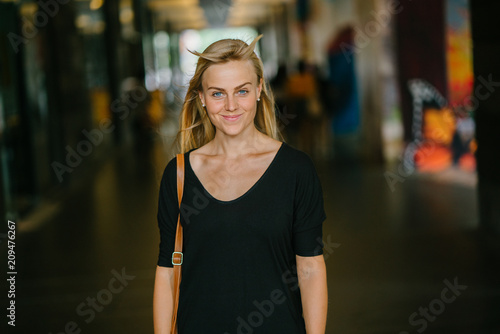 A cute blonde woman standing in the hallway is striking a confident pose in front of the camera. She is smiling and wearing a very comfortable and casual clothes while strolling in her university.