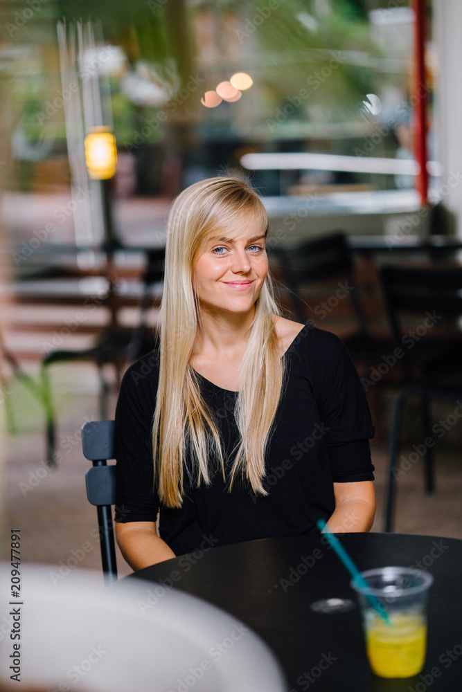 Portrait of a young blonde woman with blue eyes sitting at a cafe. She is casually and comfortably dressed. She enjoys having a drink while relaxing in a tropic city. Portrait of a young blonde woman with blue eyes sitting at a cafe. She is casually and comfortably dressed. She enjoys having a drink while relaxing in a tropic city.