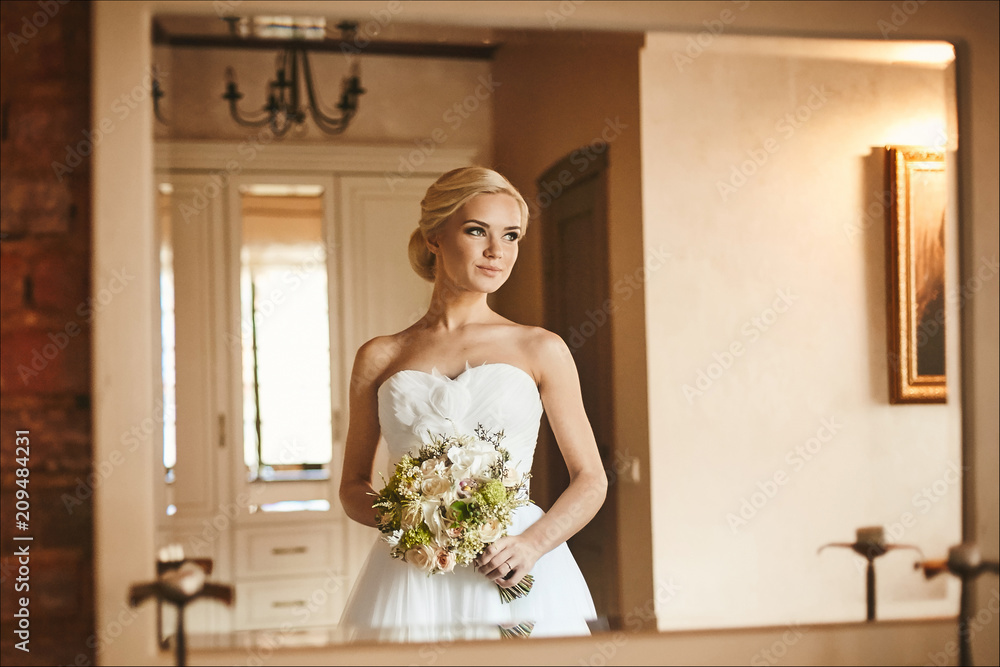 Beautiful and sensual young bride, sexy blonde model girl with charming smile and with bouquet of flowers in her hands in white dress posing in interior, wedding preparation in the morning Beautiful and sensual young bride, sexy blonde model girl with charming smile and with bouquet of flowers in her hands in white dress posing in interior, wedding preparation in the morning