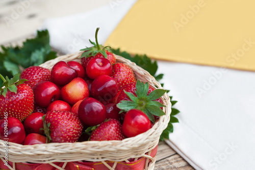 Yellow-orange fresh cherries and red ripe strawberries on a white plate, on a wooden light table with a texture, with green leaves and a white tablecloth and napkin.