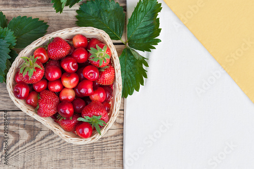 Yellow-orange fresh cherries and red ripe strawberries on a white plate, on a wooden light table with a texture, with green leaves and a white tablecloth and napkin.