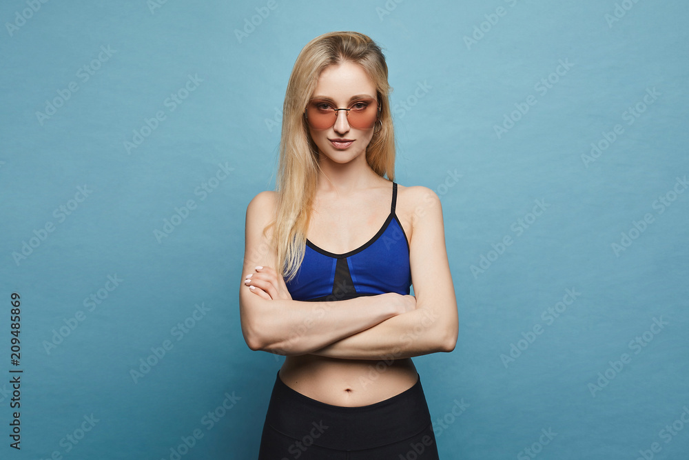 Fototapeta premium Beautiful and sexy fitness model blonde girl with perfect sporty body and makeup in sportswear and stylish sunglasses stands with folded arms and pose at blue background