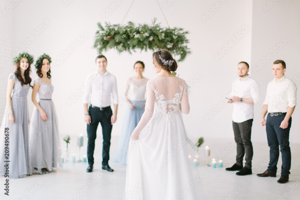 Beautiful bride in a white dress holds a wedding bouquet and is going to her groom with bridesmaids and groommen for wedding ceremony. White room is decorated with pine, flowers and blue candles. Beautiful bride in a white dress holds a wedding bouquet and is going to her groom with bridesmaids and groommen for wedding ceremony. White room is decorated with pine, flowers and blue candles.