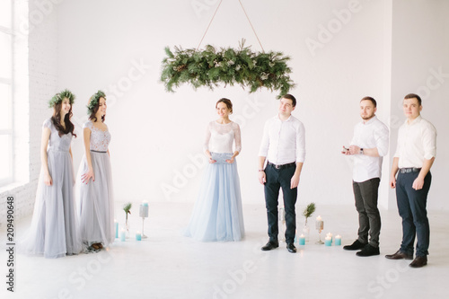 Handsome groom in white skirt and black trousers is waiting for his bride together with bridesmaids, groommen and ceremony master on the background of wedding decorations photo