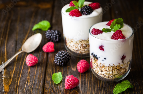 White yogurt with muesli and raspberries in glass bowl on rustic wooden background.