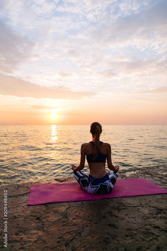 Yoga time. Healthy lifestyle concept. Back view of sportive woman meditating in lotus pose, sitting on the pier, looking at sea sunrise.