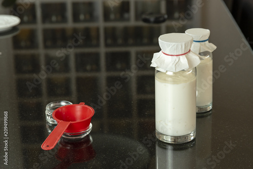 side view of a granite kitchen table top, with glass bottles with homemade kefir drink, fresh milk, red strainer, a glass plate with reflection kitchen shelf with spices in the background. photo