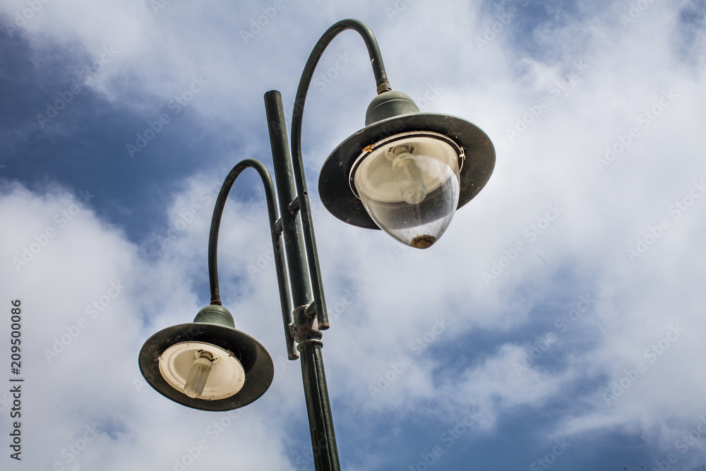 Tall old lampost with two functional bulbs. The other one is not covered and the post is painted with dark green hue. Its background is a wonderful sky blue horizon and white clouds. Tall old lampost with two functional bulbs. The other one is not covered and the post is painted with dark green hue. Its background is a wonderful sky blue horizon and white clouds.