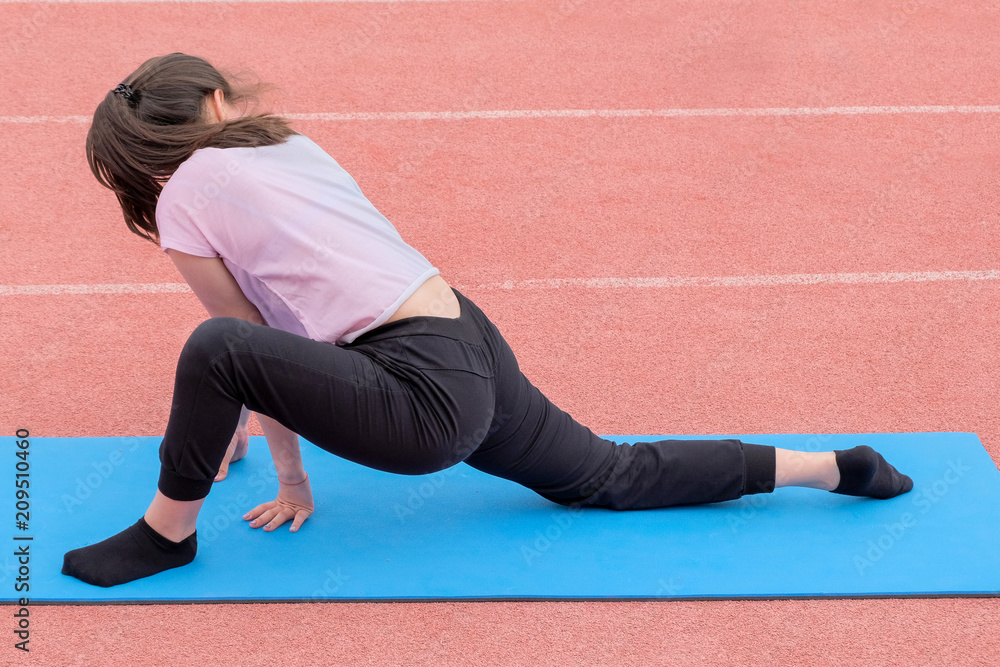 A girl in a pink T-shirt and black pants performs fitness exercises in the open air. Back view. Stretching training.