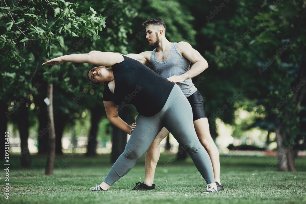 Fototapeta premium Personal trainer correct his client while doing stretching exercise. Overweight woman doing yoga with instructor support.