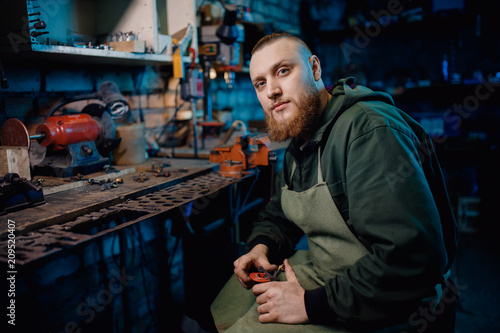 Man with beard in overalls makes details of iron and wood in garage workshop, handmade production. concept is professional jeweler.