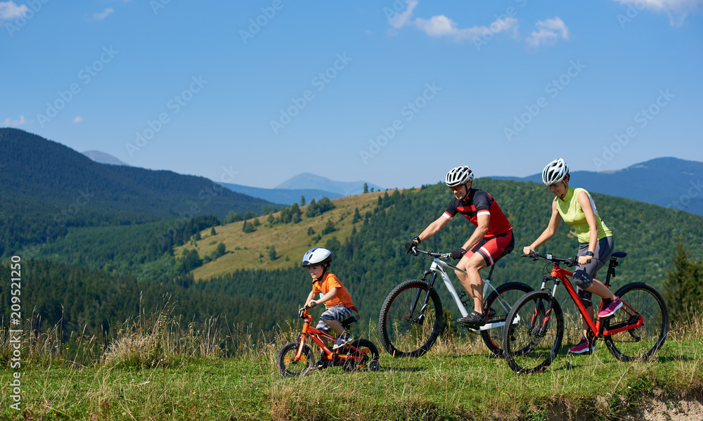 Young modern family tourists bikers, mom, dad and child riding on bicycles on grassy hill. Carpathian mountains, blue summer sky on background. Active lifestyle, traveling and happy relations concept.