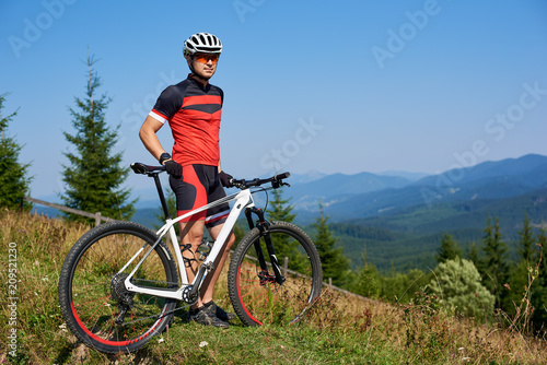 Full length portrait of athletic professional cyclist standing on top of hill, enjoying view of distant Carpathian mountains on blue summer sky background. Active lifestyle and outdoor sport concept.