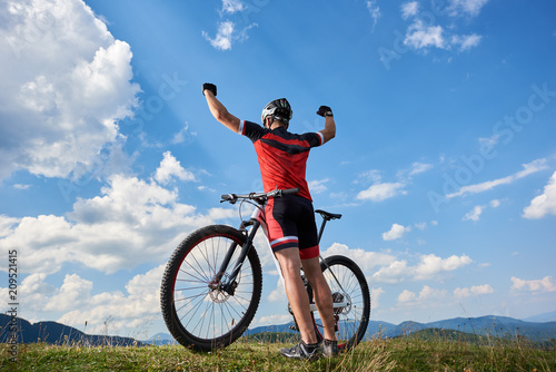 Back view of athletic professional sportsman cyclist standing with bike on top of hill, rasing hands, against blue sky with clouds on summer sunny day. Outdoor sport, success and life goals concept