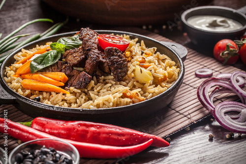 The concept of eastern, Uzbek cuisine. Pilaf, plov in a black cast-iron frying pan on a wooden table, next to lie ingredients, onions, tomatoes, parsley, pepper. Central Asian cuisine.