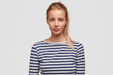 Horizontal shot of pretty female with light hair combed in pony tail, wears sailor sweater, listens attentively interlocutor, poses against white background. Confident woman looks directly at camera