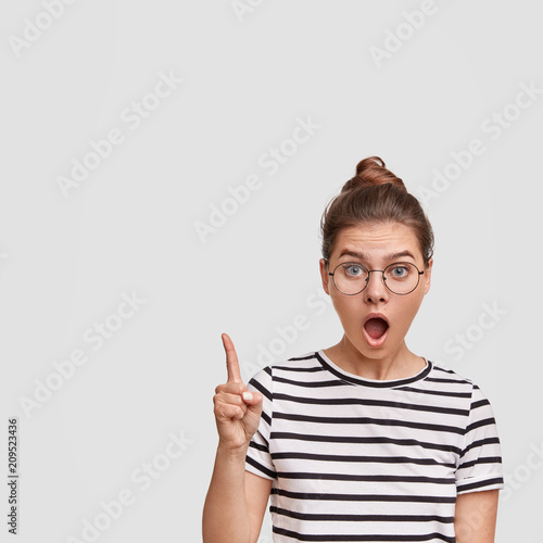 Puzzled stupefied female in glasses, being shocked as her neigbour flooded apartment again, points up with fore finger, lifts eyebrows in surprisement, keeps jaw dropped, isolated on white wall photo
