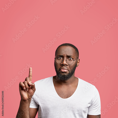 Indoor shot of plump dark skinned young male looks in bewilderment, indicates with fore finger upwards, has puzzled expression, shows blank space for your advertisement, dressed in white t shirt photo