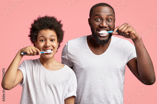 Positve dark skinned son and father brush teeth, keep mouthes wide opened, have satisfied expressions, take care of health, dressed casually, isolated over pink background. Morning routine concept