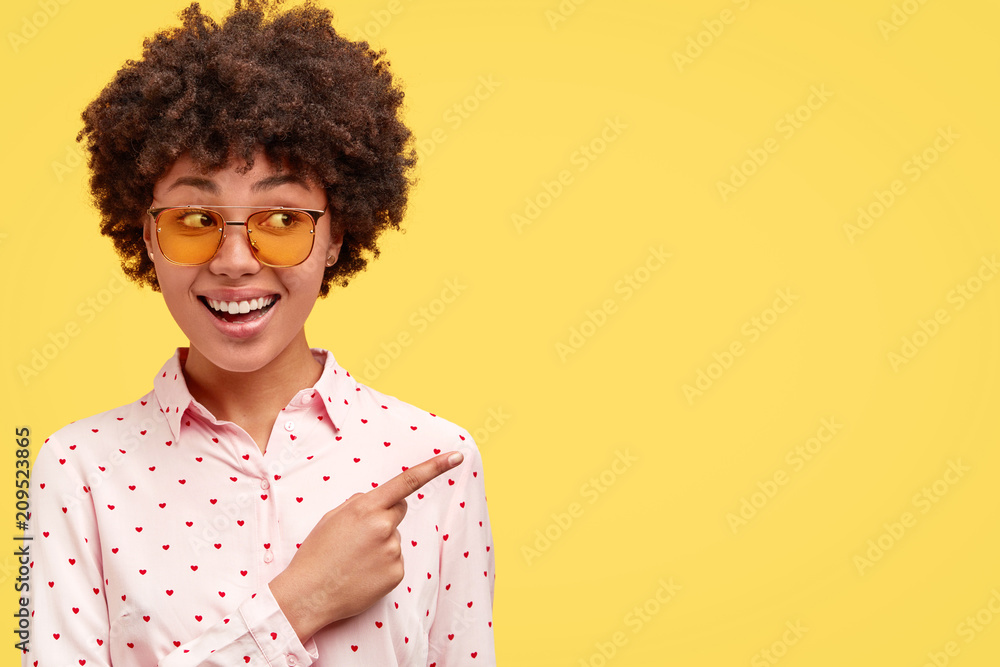 Positive African American female with curly hair, shows you place for entertainment or good pub, inidicates aside with fore finger, invites to go there, poses on yellow background. Ethnicity concept Positive African American female with curly hair, shows you place for entertainment or good pub, inidicates aside with fore finger, invites to go there, poses on yellow background. Ethnicity concept
