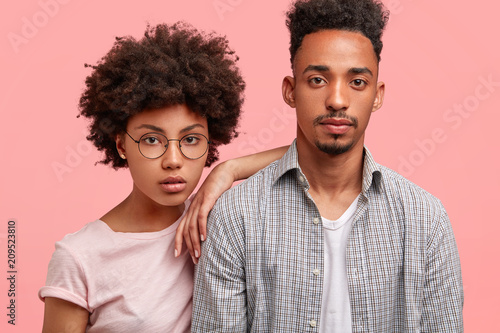 Studio shot of serious African American couple in love come to tour agency as want choose some travel during vacation, look attentively at price list, isolated over pink background. Friendship concept