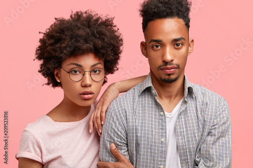 Lovely African American couple stand close to each other, look seriously at camera, have confident expressions, dressed in casual outfit, pose together against pink background. Companionship concept