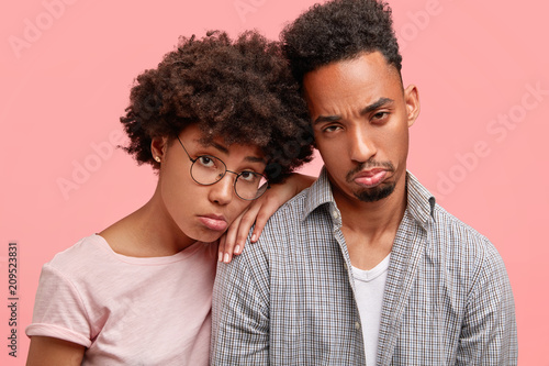 Unhappy African American couple have depressed and sad expressions, have financial problem, feel desperate, purse lips, need support and help, isolated over pink background. Negative emotions
