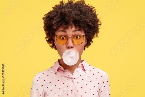 Fotografie African American female blows bubble from chewing gum, has amazed expression, dressed in fashionable clothes and shades, isolated over yellow background